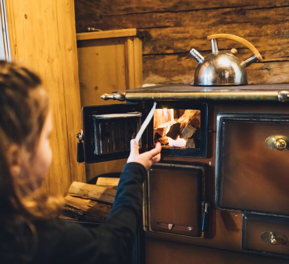Ein rustikaler Holzofen in einer ländlichen Umgebung wird von einer Person befeuert, die Brennholz in die Ofenkammer legt. Ein alter Kochtopf auf dem Ofen deutet auf die Nutzung als Heizquelle und zum Kochen hin. | © Urlaub am Bauernhof Salzburg / Daniel Gollner 