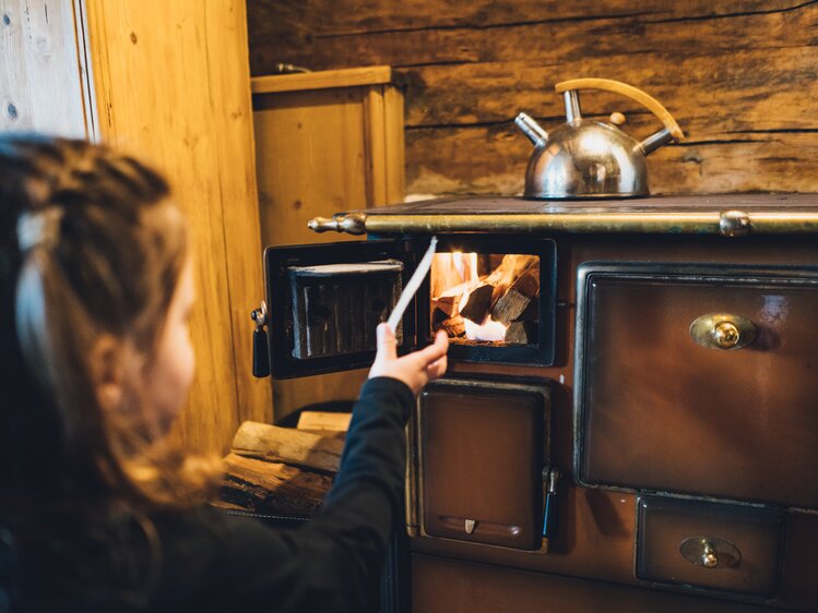 Ein rustikaler Holzofen in einer ländlichen Umgebung wird von einer Person befeuert, die Brennholz in die Ofenkammer legt. Ein alter Kochtopf auf dem Ofen deutet auf die Nutzung als Heizquelle und zum Kochen hin. | © Urlaub am Bauernhof Salzburg / Daniel Gollner 