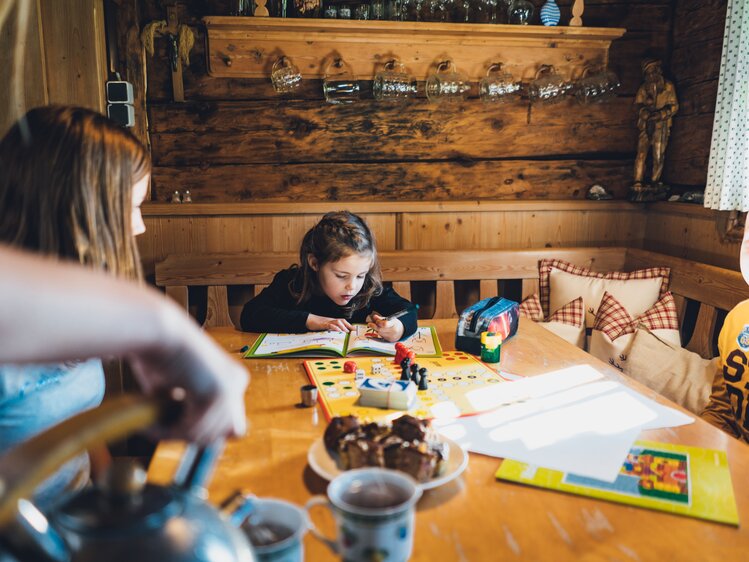Kinder spielen und lesen am Holztisch in einem gemütlichen Heim, umgeben von rustikalen Dekorationen. Eine Tasse und ein Teller mit Süßigkeiten ergänzen die Szene. | © Urlaub am Bauernhof Salzburg / Daniel Gollner 