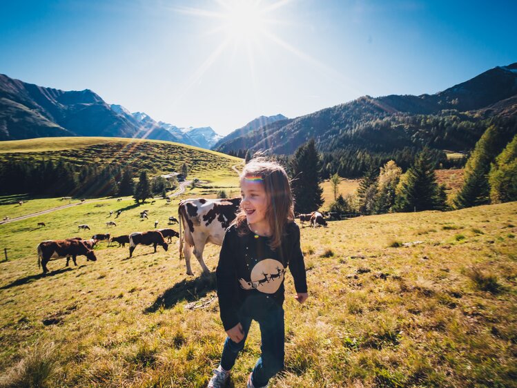Fröhliches junges Mädchen in schwarzer Kleidung, das inmitten einer malerischen Berglandschaft mit Kühen steht. Die Sonne scheint, und die Bergkette zeichnet sich im Hintergrund ab. | © Urlaub am Bauernhof Salzburg / Daniel Gollner 