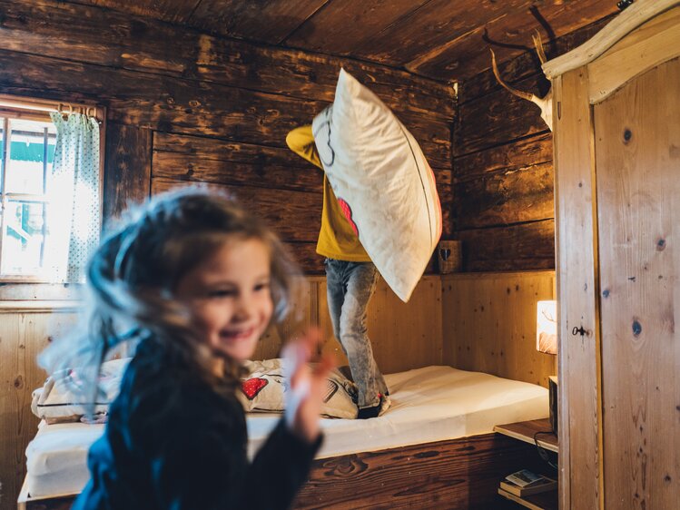 Kleines Mädchen lächelt fröhlich in einer gemütlichen Holzhütte, ein Erwachsener wirft gerade ein Kissen auf eine Bettstelle. | © Urlaub am Bauernhof Salzburg / Daniel Gollner 