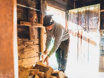 Eine Person in einem karierten Hemd und Kappe bearbeitet Holzbretter in einer rustikalen Werkstatt mit Holzwänden und -konstruktionen. Im Hintergrund sind verschiedene Werkzeuge und Dekorationen zu sehen. | © Urlaub am Bauernhof Salzburg / Daniel Gollner 