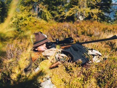 Eine Person in Tarnkleidung und Hut liegt mit einem Gewehr bewaffnet auf dem Boden, umgeben von herbstlicher Vegetation. | © Urlaub am Bauernhof Salzburg / Daniel Gollner 