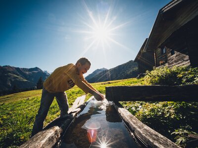 Hände waschen im Brunnen | © Urlaub am Bauernhof Salzburg / Daniel Gollner Eine Person in gelbem Oberteil, die an einem hölzernen Brunnen auf einer sonnendurchfluteten Wiese beim Wasserschöpfen zu sehen ist. Im Hintergrund erheben sich bewaldete Berge unter einem strahlend blauen Himmel. | © Urlaub am Bauernhof Salzburg / Daniel Gollner