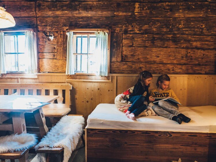 Zwei Kinder sitzen auf einem Holzbett in einer traditionell eingerichteten Holzhütte. Sie lesen und genießen die gemütliche Umgebung mit rustikalen Möbeln und Fenstern. | © Urlaub am Bauernhof Salzburg / Daniel Gollner 
