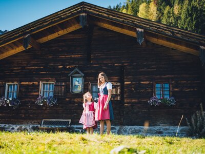 Eine Frau und ein Mädchen in traditioneller bayerischer Tracht vor einer rustikalen Holzhütte mit Blumenbeeten und umgeben von berghafter Landschaft. | © Urlaub am Bauernhof Salzburg / Daniel Gollner 