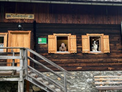 Altes Holzhaus mit Fensterläden, Aufbauten aus Holzkisten und Leiter angelehnt an der Fassade, umgeben von Grün. | © Urlaub auf der Alm / Julia Winkler