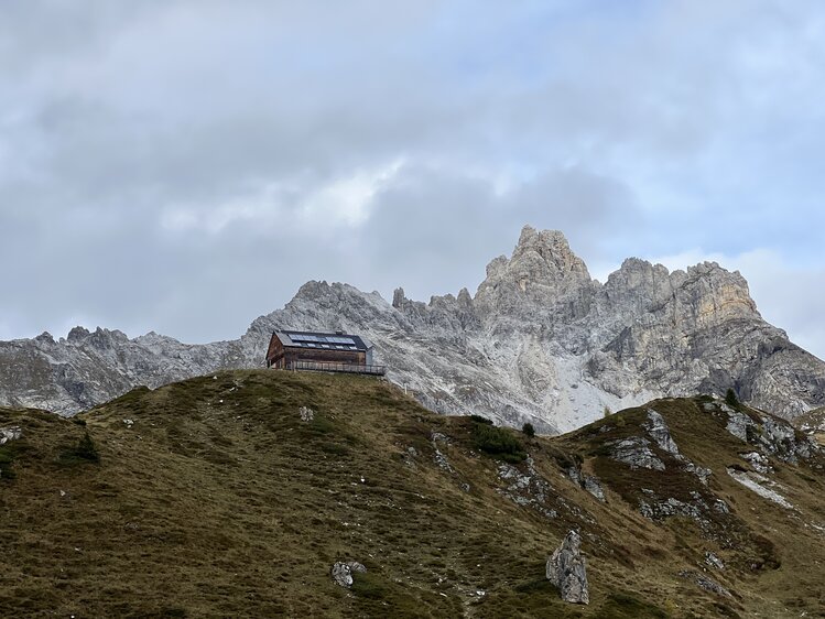 Eine kleine Hütte auf einem Hügel, umgeben von majestätischen, schneebedeckten Bergen in einer nebligen Landschaft. | © Urlaub auf der Alm / Julia Winkler