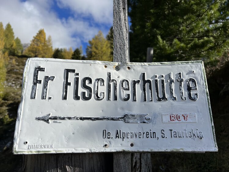Ein Wegweiser in der Natur mit deutscher Aufschrift "Fr. Fischerhutte" und der Information "De. Alpenverein, S. Tauriskie", umgeben von buntem Herbstlaub und blauem Himmel. | © Urlaub auf der Alm / Julia Winkler