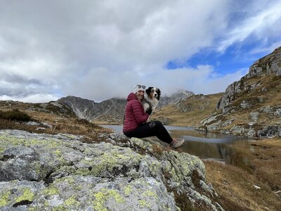 Eine Person sitzt auf einem felsigen Ufer neben einem Bergsee, begleitet von einem Hund. Der Himmel ist bewölkt mit Blau und Grau, die Berge ragen dahinter auf. Eine ruhige, bergige Landschaft. | © Urlaub auf der Alm / Julia Winkler