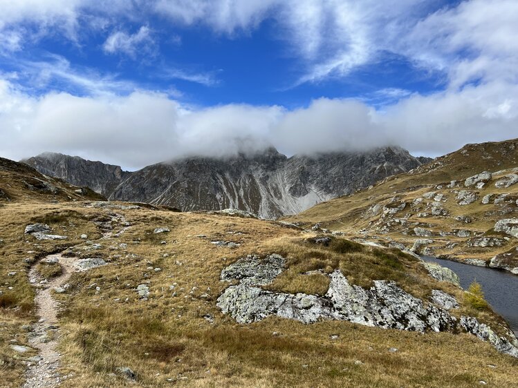 Berglandschaft mit schneebedeckten Gipfeln, eingehüllt in Wolken, umgeben von felsiger Umgebung und Grasland. Ein einfacher Weg schlängelt sich durch die Landschaft. | © Urlaub auf der Alm / Julia Winkler