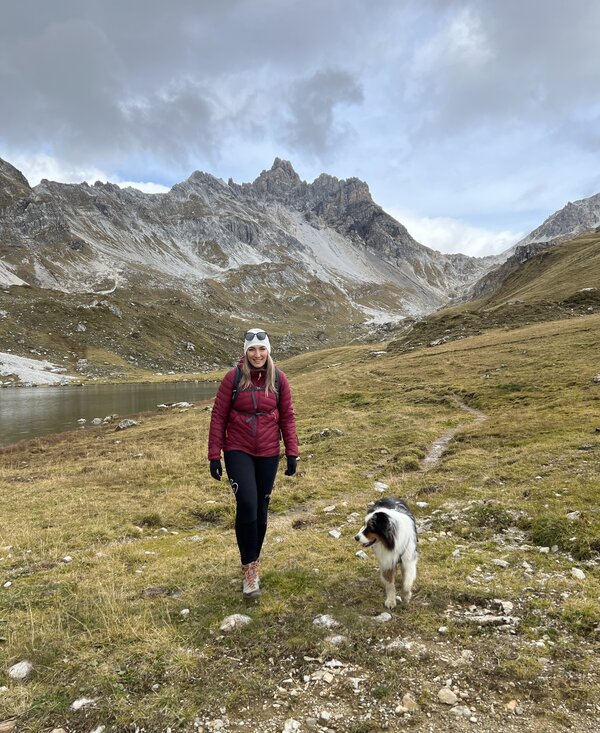 Eine Frau in einer roten Jacke spaziert mit ihrem Hund an einem See in einer bergigen Landschaft. Im Hintergrund sind felsige Berge mit schneebedeckten Spitzen zu sehen. | © Urlaub auf der Alm / Julia Winkler