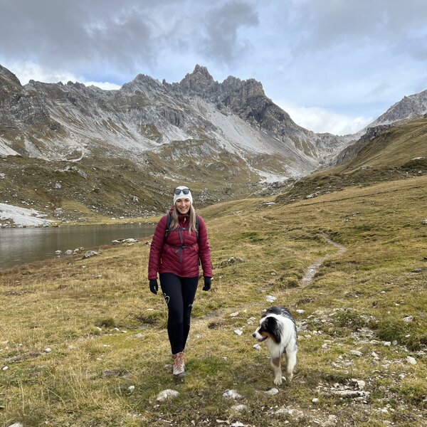 Eine Frau in einer roten Jacke spaziert mit ihrem Hund an einem See in einer bergigen Landschaft. Im Hintergrund sind felsige Berge mit schneebedeckten Spitzen zu sehen. | © Urlaub auf der Alm / Julia Winkler
