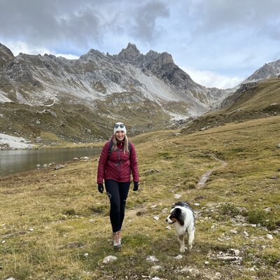 Eine Frau in einer roten Jacke spaziert mit ihrem Hund an einem See in einer bergigen Landschaft. Im Hintergrund sind felsige Berge mit schneebedeckten Spitzen zu sehen. | © Urlaub auf der Alm / Julia Winkler