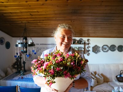 Gastgeberin Theresia Bacher mit einem Strauß Almrausch auf der Dürsteinalm | © Urlaub am Bauernhof / Daniel Gollner Eine fröhliche ältere Frau hält einen Blumenstrauß in der Hand und lächelt in einer gemütlichen, häuslichen Umgebung mit hölzerner Decke und gerahmten Dekorationen an den Wänden. | © Urlaub am Bauernhof / Daniel Gollner