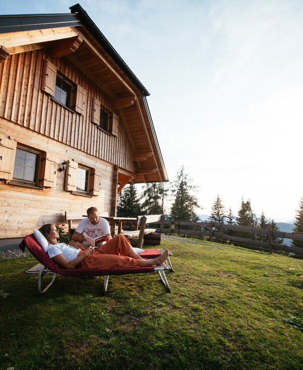 Eine Holzhütte auf einer Wiese mit zwei Personen, die auf Liegen in der Sonne sitzen und den Ausblick auf die Berglandschaft genießen. | © Urlaub am Bauernhof Steiermark / Daniel Gollner