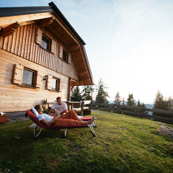 Eine Holzhütte auf einer Wiese mit zwei Personen, die auf Liegen in der Sonne sitzen und den Ausblick auf die Berglandschaft genießen. | © Urlaub am Bauernhof Steiermark / Daniel Gollner