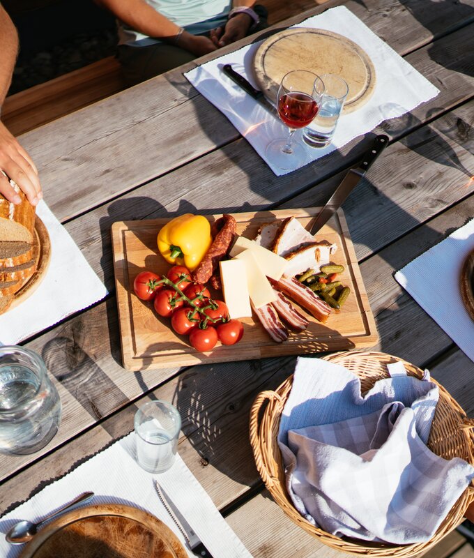 Gemütliches Holztisch-Picknick im Freien mit Brot, Käse, Wurst, Oliven und Gläsern für Wein oder Schnaps. Frische Früchte und Gemüse sowie Besteck und Servietten vervollständigen die Tischausstattung. | © Urlaub am Bauernhof Steiermark / Daniel Gollner