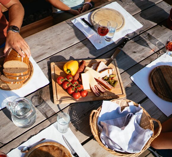 Gemütliches Holztisch-Picknick im Freien mit Brot, Käse, Wurst, Oliven und Gläsern für Wein oder Schnaps. Frische Früchte und Gemüse sowie Besteck und Servietten vervollständigen die Tischausstattung. | © Urlaub am Bauernhof Steiermark / Daniel Gollner