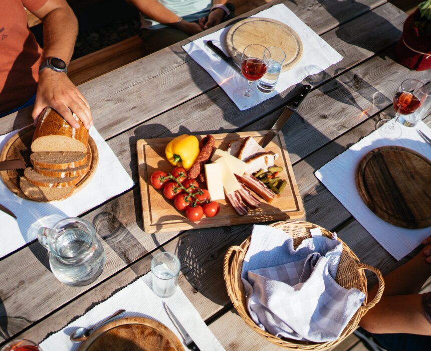 Gemütliches Holztisch-Picknick im Freien mit Brot, Käse, Wurst, Oliven und Gläsern für Wein oder Schnaps. Frische Früchte und Gemüse sowie Besteck und Servietten vervollständigen die Tischausstattung. | © Urlaub am Bauernhof Steiermark / Daniel Gollner