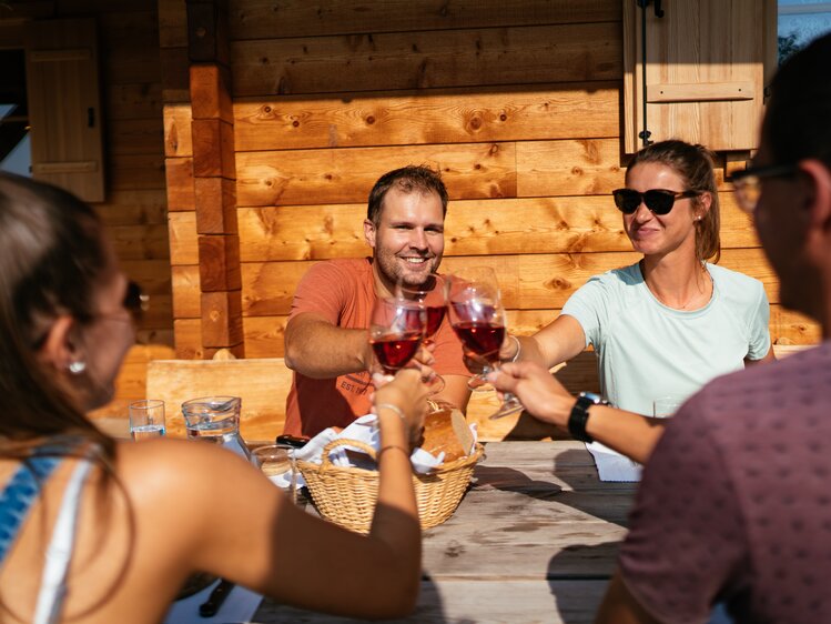 Eine Gruppe von Erwachsenen sitzt an einem Holztisch auf einer Veranda und genießt Wein. Sie stoßen miteinander an und lachen, was auf eine gemütliche, gesellige Atmosphäre hindeutet. | © Urlaub am Bauernhof Steiermark / Daniel Gollner 