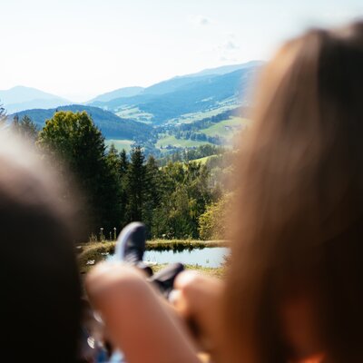 Weite, bergige Landschaft mit Wäldern, einem See und einer Person am Berg, die den Blick über die Umgebung genießt. | © Urlaub am Bauernhof Steiermark / Daniel Gollner
