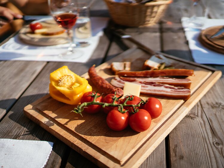 Holzbrett mit Tomaten, Paprika und Speck, umgeben von Besteck, Glas und weiteren Lebensmitteln auf einem hölzernen Tisch. | © Urlaub am Bauernhof Steiermark / Daniel Gollner 