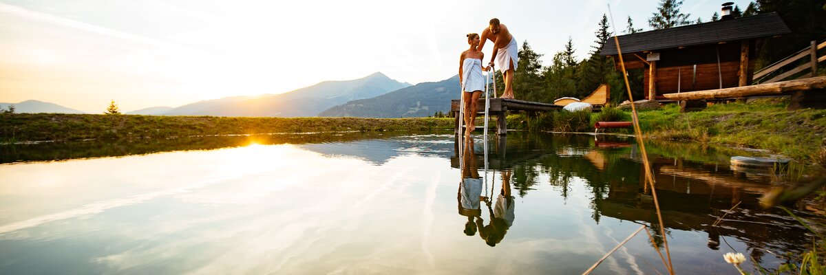Ein friedliches Paar genießt den Blick auf den See mit einer Holzstegplattform im Vordergrund und einer einladenden Landschaft im Hintergrund bei Sonnenuntergang. | © Urlaub am Bauernhof Steiermark / Daniel Gollner
