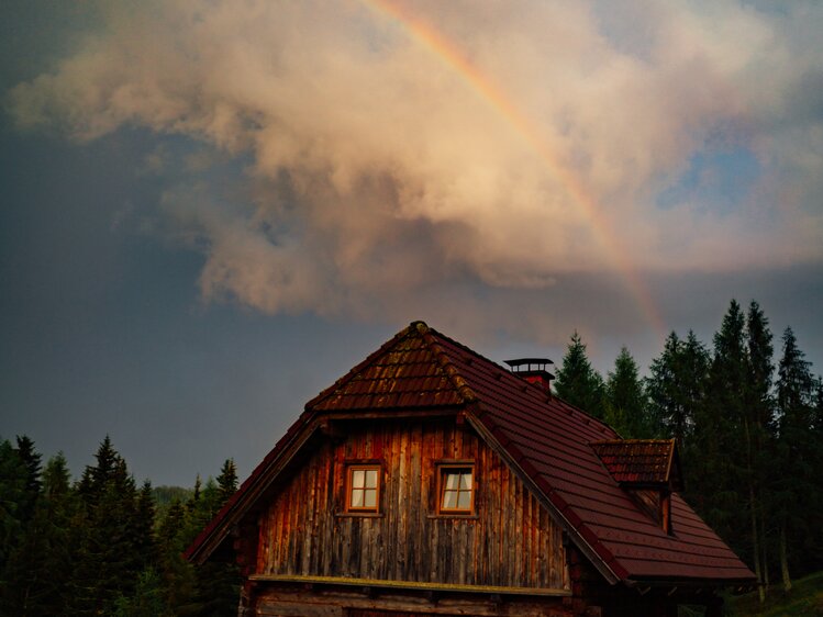 Ein traditionelles Holzhaus mit einem Strohdach und beleuchteten Fenstern, umgeben von Bäumen und einem Regenbogen am Himmel. Die Szene strahlt eine gemütliche und friedliche Atmosphäre aus. | © Urlaub am Bauernhof Steiermark / Daniel Gollner 