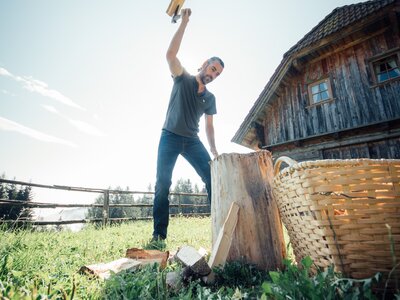 Ein Mann in blauem T-Shirt und Jeans steht vor einer alten Holzhütte, hebt eine Axt hoch und scheint Holz zu hacken. Im Vordergrund ist ein mit Gras bewachsener Hügel zu sehen, im Hintergrund eine ländliche Landschaft. | © Urlaub am Bauernhof Steiermark / Daniel Gollner 
