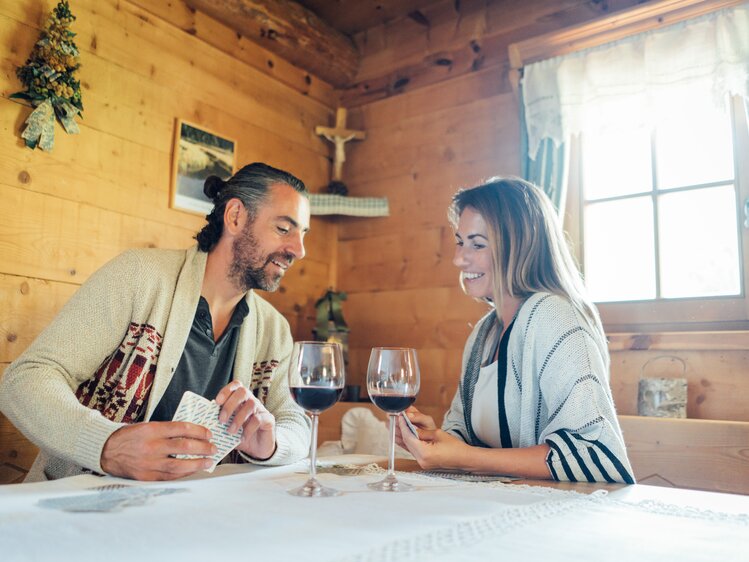 Ein Paar genießt Rotwein in einer gemütlichen Holzhütte, geschmückt mit Weihnachtsdekorationen und einem Kruzifix an der Wand. Sie lächeln und scheinen eine entspannte, romantische Atmosphäre zu teilen. | © Urlaub am Bauernhof Steiermark / Daniel Gollner 