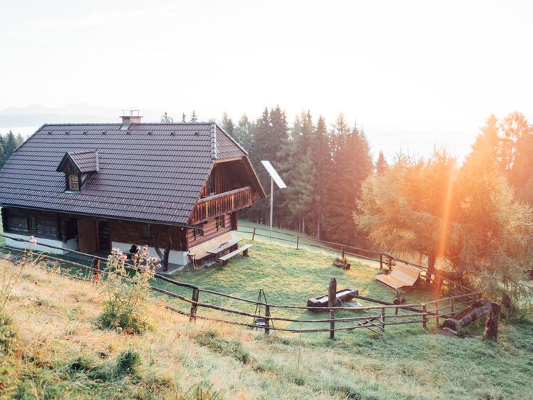 Malerische Holzhütte umgeben von Bergwald, Wiesen und Sonnenlicht. Traditioneller Baustil mit Dachsparren und Balkon, Umzäunung und Aufgang. Friedliche, herbstliche Szenerie. | © Urlaub am Bauernhof Steiermark / Daniel Gollner 
