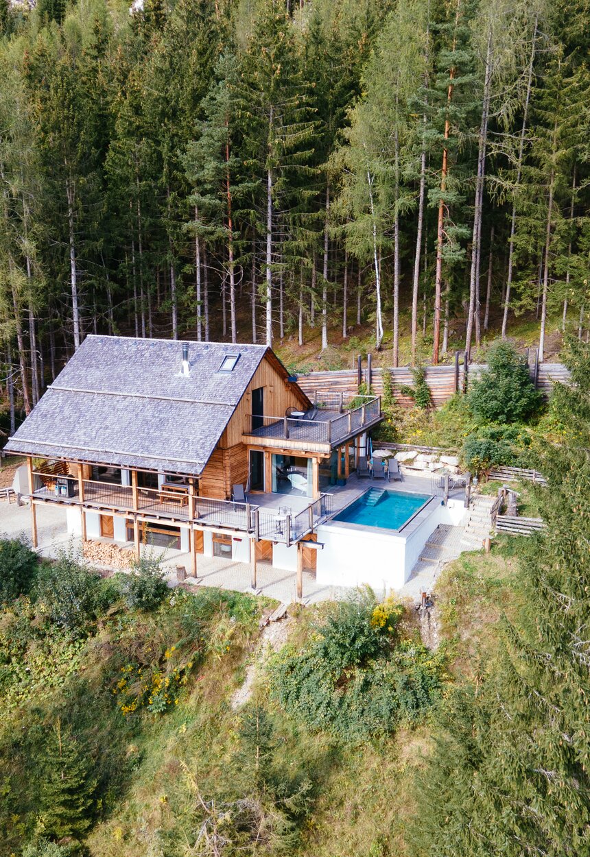 Holzhaus mit Schwimmbad inmitten eines Waldgebiets, umgeben von hohen Bäumen und grüner Vegetation. Das Gebäude verfügt über einen teilweise überdachten Balkon oder Terrasse mit Blick auf den Pool. | © Urlaub am Bauernhof Steiermark / Daniel Gollner