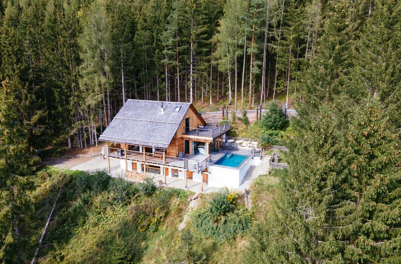 Holzhaus mit Schwimmbad inmitten eines Waldgebiets, umgeben von hohen Bäumen und grüner Vegetation. Das Gebäude verfügt über einen teilweise überdachten Balkon oder Terrasse mit Blick auf den Pool. | © Urlaub am Bauernhof Steiermark / Daniel Gollner