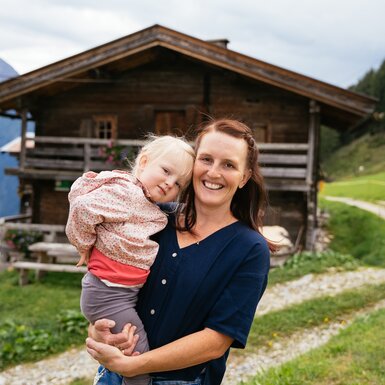 Lächelnde Mutter hält ihr kleines Mädchen vor einem rustikalen Holzhaus in einer idyllischen Berglandschaft. | © Urlaub am Bauernhof Tirol / Daniel Gollner 