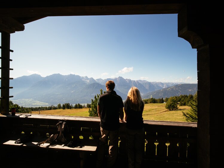 Ein Paar genießt den Ausblick von einem Balkon aus auf eine malerische Gebirgslandschaft mit bewaldeten Hügeln und einem klaren blauen Himmel. | © Urlaub am Bauernhof Tirol / Daniel Gollner 
