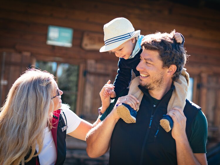 Eine junge Familie genießt einen Spaziergang an einem Holzhaus. Der Vater trägt den Sohn auf den Schultern, während die Mutter neben ihnen läuft. Alle lächeln und scheinen eine fröhliche Zeit miteinander zu verbringen. | © Urlaub am Bauernhof Tirol / Daniel Gollner 