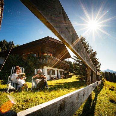 Zwei entspannte Personen auf einer Wiese vor einer traditionellen Holzhütte sitzen, flankiert von Bergen und blauem Himmel mit strahlendem Sonnenlicht. | © Urlaub am Bauernhof Tirol / Daniel Gollner 