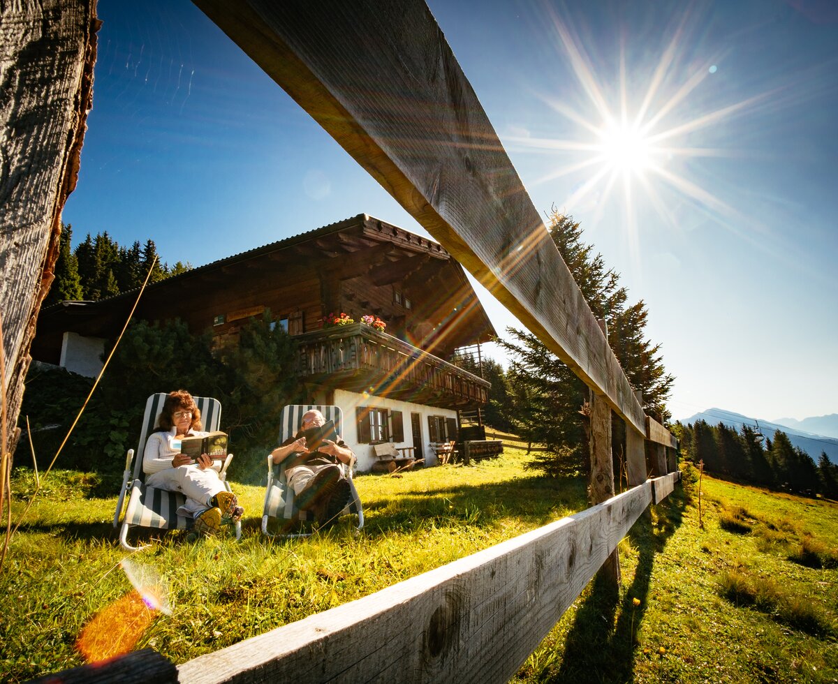 Grosseltern entspannen vor der Hütte | © Urlaub am Bauernhof Tirol / Daniel Gollner Zwei entspannte Personen auf einer Wiese vor einer traditionellen Holzhütte sitzen, flankiert von Bergen und blauem Himmel mit strahlendem Sonnenlicht. | © Urlaub am Bauernhof Tirol / Daniel Gollner