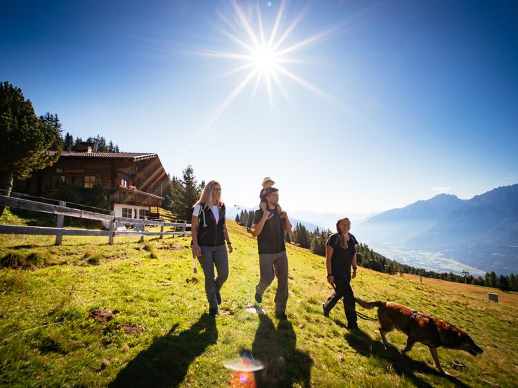 Wanderer genießen einen sonnigen Tag in den Bergen, umgeben von Wiesen, Wäldern und einem Berghaus. Ein Hund begleitet die Gruppe auf ihrem Weg. | © Urlaub am Bauernhof Tirol / Daniel Gollner 
