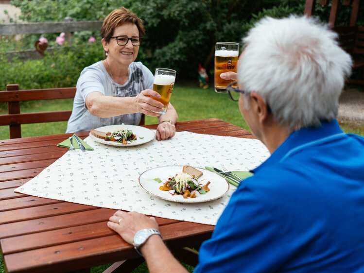 Ein Frau und ein Mann sitzen im Garten und stoßen mit einem Bier an. Am Tisch steht ein schön angerichtetes Essen. | © Urlaub am Bauernhof Kärnten/ Angelina Tschische