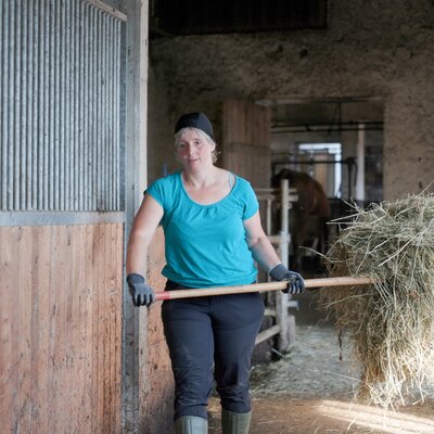 Frau in landwirtschaftlicher Halle trägt Heu auf einer Plane auf einem Holzgestell.