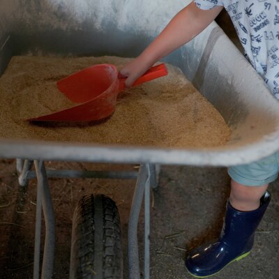 Ein Kind schaufelt Sand mit einem roten Schaufel in einer Hangkarre, trägt ein T-Shirt mit verschiedenen Symbolen und Shorts.