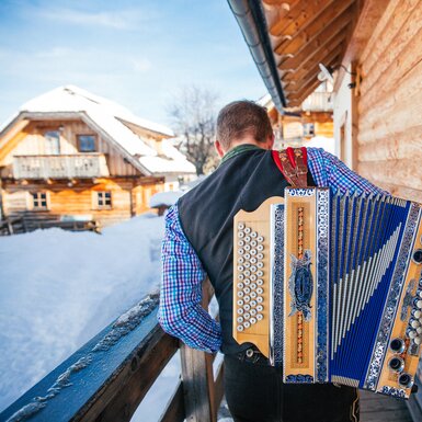 Musiker geht mit einer Steirischen Ziehharmonika am Rücken an einer Hütte vorbei | © Urlaub am Bauernhof Oberösterreich / Pascal Baronit