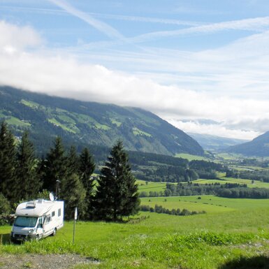 Grüne Wiesen, bewaldete Hügel und Berge, die sich in der Ferne erstrecken. Wohnmobile stehen auf einem Campingplatz umgeben von der malerischen Landschaft.