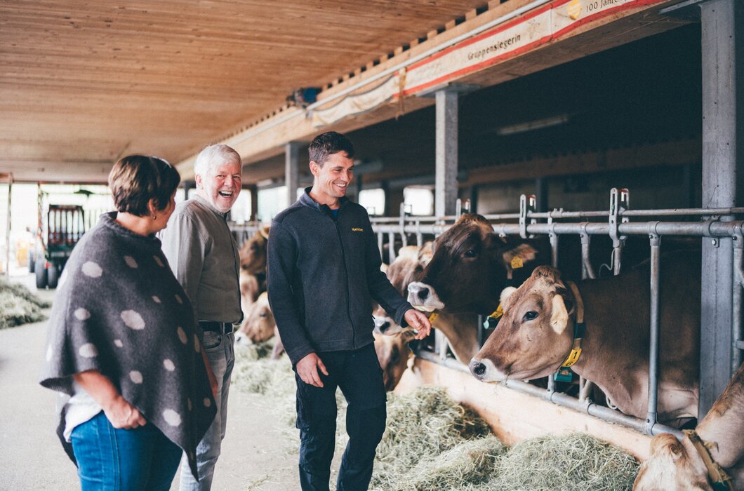 Drei Personen lächeln in einem Kuhstall voller Kühe. Die Umgebung ist mit Holz und Metallstangen ausgestattet, was auf einen landwirtschaftlichen Betrieb hinweist. | © Urlaub am Bauernhof / Daniel Gollner
