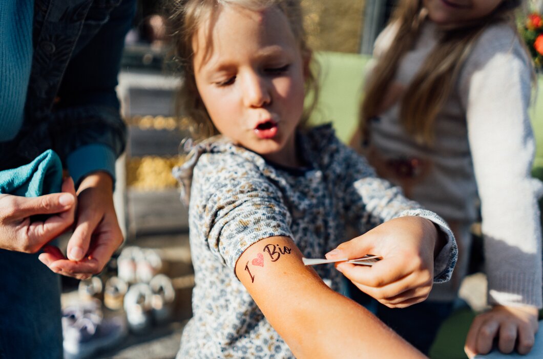 Ein Mädchen erhält eine Tätowierung auf dem Arm, während eine erwachsene Person hilft. Es scheint eine freundliche, kreative Aktivität zu sein.