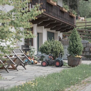 Ein idyllisches Berghäuschen mit Holzbalkonen, umgeben von Bäumen und grüner Natur. Auf der Terrasse stehen Holzmöbel und ein Spielzeugtraktor, was eine gemütliche Atmosphäre vermittelt. | © Urlaub am Bauernhof / Daniel Gollner