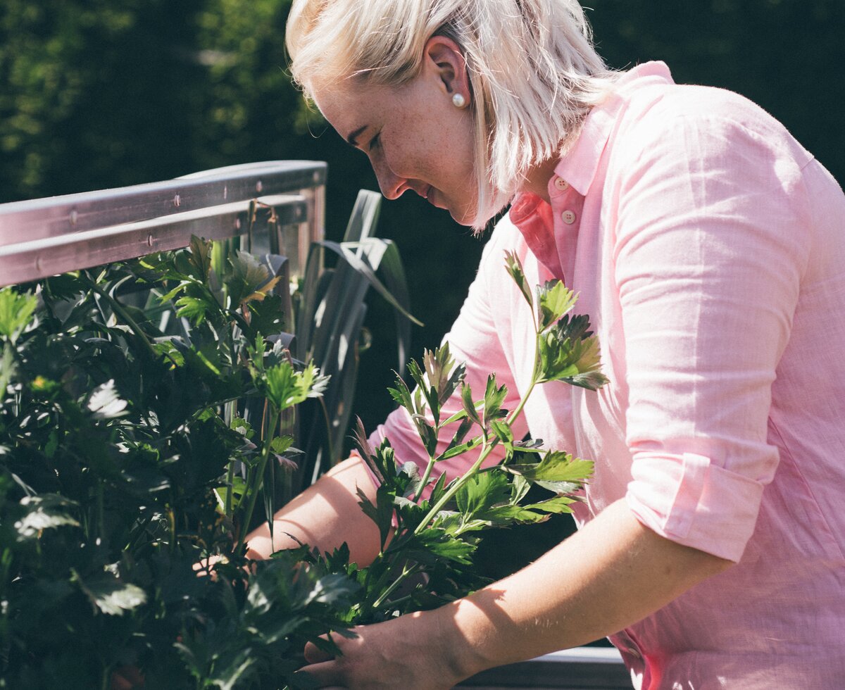 Eine Person in rosa Kleidung, die Blumen in einem Blumenkasten pflegt. Die Umgebung ist grün und natürlich, vermutlich ein Garten oder eine Gartenanlage. Die Szene wirkt friedlich und kontemplativ. | © Urlaub am Bauernhof / Daniel Gollner