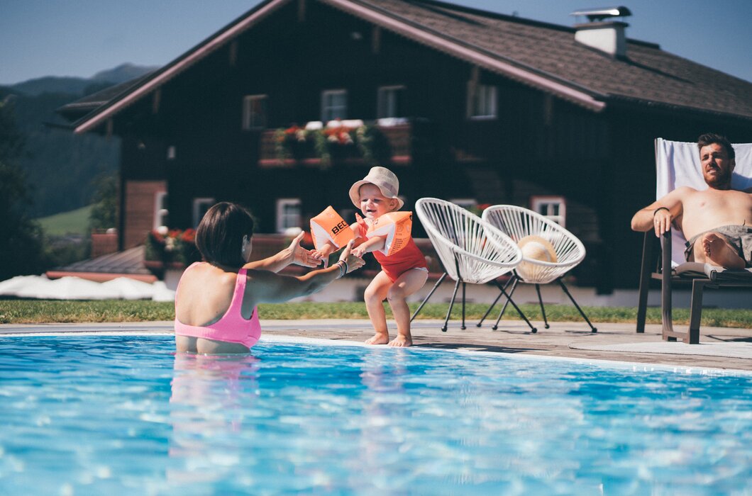 Familie am Pool | © Urlaub am Bauernhof / Daniel Gollner Eine Frau im Badeanzug begrüßt ein Baby am Beckenrand, während ein Mann in Liegestühlen sitzt und beobachtet. Ein Haus mit Blumenkästen im Hintergrund. | © Urlaub am Bauernhof / Daniel Gollner